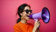© miss irine - Woman in orange top holds megaphone. Wears sunglasses, gold earrings. Woman enthusiastically speaking into megaphone against vibrant pink background. Possible scenario social media ad, online