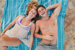 © Jose Calsina - Joyful Young Couple Relaxing on a Colorful Beach Towel Under the Sun Smiling and Enjoying a Blissful Summer Day Together at the Shoreline. Two young people laying on the sand and looking at camera.