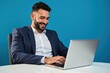 © velikiyzayats - Professional Man in a Suit Working on a Laptop in a Modern Office Setting with Blue Background, Smiling and Engaged in Productive Activity