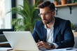 © velikiyzayats - Focused Young Adult Male Professional in a Stylish Office Setting Working on a Laptop Surrounded by Greenery and Modern Decor