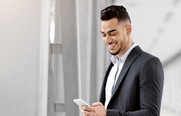 Wall Mural - Business Trip. Handsome Young Arab Businessman Using Smartphone In Airport Terminal, Smiling Middle Eastern Entrepreneur Holding Passport With Tickets, Waiting For Flight Boarding, Free Space