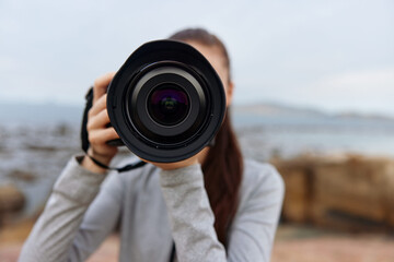 Young female photographer holding a camera outdoors, showcasing a serene expression and casual attire, with a natural coastal background and soft color palette