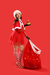 Wall Mural - Pretty young woman in Santa costume with tasty burger and bag of gifts on red background