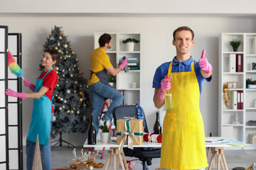 Canvas Print - Male janitor with detergent showing thumb-up against his team cleaning in office after New Year party