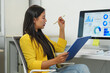 © Phimwilai - Focused businesswoman in a modern office analyzing digital data on a computer screen, holding a pen and clipboard. Engaged in reviewing statistics and graphs for business insights