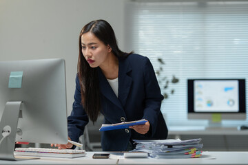 Focused asian businesswoman working diligently at a computer while holding a clipboard, standing at a modern desk surrounded by a stack of important documents in a corporate office