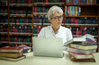 © APchanel - Elderly woman sitting in a library, combining traditional and modern approaches to learning or research by book and laptop computer sense of purpose dedication to personal grown concept.