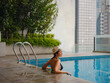 © YURII Seleznov - Young asian woman relaxing by pool at Kuala Lumpur hotel with view of surrounding skyscrapers, enjoying leisure time in vibrant urban setting.