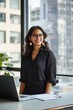 © felix_brönnimann - Businesswoman in modern office smiling at the camera while working on a laptop. Casual attire, creative workspace, and urban skyline in background. Professional lifestyle.