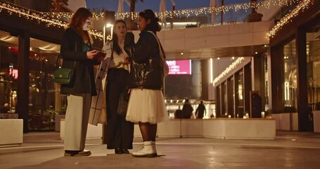 Canvas Print - Three diverse businesswomen collaborate on a project outdoors at night, discussing ideas and strategies in a lively urban setting.