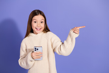 Wall Mural - Smiling young girl in casual sweater holding phone and pointing, vibrant violet background, advertising or promoting concept.