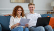 © Multi7n - Young Couple Sitting on Blue Couch Reviewing Paper with Laptop in Cozy Living Room