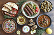 © Myimages - Middle eastern, arabic or mediterranean dinner table with grilled lamb kebab, chicken skewers with roasted vegetables and appetizers variety serving on wooden outdoor table. Overhead view.
