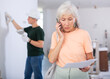 © JackF - Portrait of emotional mature woman displeased with quality of repair work having phone call in apartment under reconstruction, teenager maintenance worker standing behind
