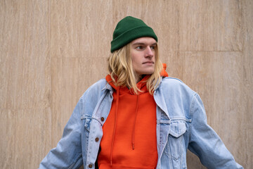 Blonde long-haired hipster guy in colorful trendy stylish outfit, posing, leaning against wall in city. Carefree fashionable male in orange hoodie and jean, representative of generation z