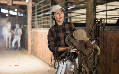 Wall Mural - Confident smiling Asian female stable keeper carrying horse saddle and harness for horseback ride