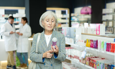 Wall Mural - Attentive mature woman choosing convenient lubricant standing next to shelves in chemist's shop