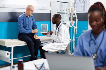 Wall Mural - Caucasian elderly man seated on hospital bed as african american doctor uses tablet to explain his medical results. Male health specialist holds digital device while consulting with senior patient.