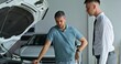 © m-art - A car dealership manager talks to a customer standing next to a car with the hood open in the showroom
