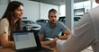 © m-art - A man signs a car purchase agreement at a car dealership. The manager is sitting at his desk in front of an open laptop