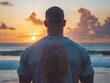 © somchai20162516 - A man with a large back tattoo, wearing a fitted shirt, standing in front of a beach at sunset, with his tattoo visible as he gazes into the distance.