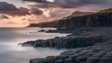 Poster - Serene coastal landscape at sunset featuring dark granite rock formations in the foreground with soft waves and cloud-streaked sky in the background.