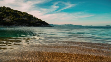 Aufkleber - Serene coastal landscape with clear turquoise water and gentle ripples in the foreground, lush green hillside on the left under a blue sky