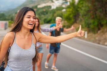 Wall Mural - cheerful interracial friends standing on road showing hitchhiking gestures. tourism traveling