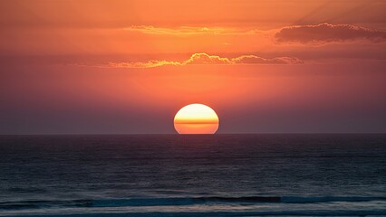 Poster - Serene sunset over the ocean featuring a vibrant orange sun against a deep blue sky with soft clouds reflecting warm hues and tranquil waves below.