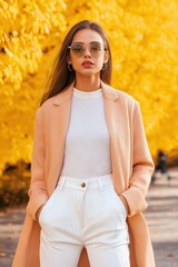 Poster - A woman wearing a white shirt and tan coat stands in front of a tree with yellow leaves. She is wearing sunglasses and has her hands in her pockets. Concept of casual elegance and a relaxed atmosphere