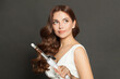 © artmim - Attractive woman with curly hairstyle using hot curling tongs iron, indoor fashion portrait. Coiffure, beauty salon and hair styling concept