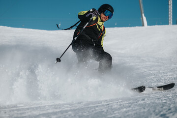 Wall Mural - An experienced skier passionately descends a snow-covered slope, surrounded by a spray of snow and clear skies. Their focus and skill showcase the excitement and beauty of winter sports.