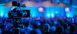 © Emvats - A professional video camera on a tripod records a live event in a dimly lit venue. The operator adjusts the camera, capturing the performance under blue stage lighting.