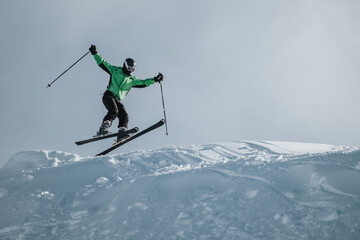 A skier wearing green and black performs an airborne jump above a snowy hilltop under clear skies. The dynamic action and pristine landscape showcase the thrill of skiing and winter sports.