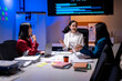 © Wasana - Three women are sitting at a table in a room with a large monitor on the wall