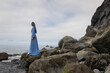 © Milou Dirks - art portrait of woman standing on the rocks near beach and ocean looking over the sea wearing long blue dress