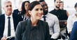 © peopleimages.com - Woman, listening and smile in crowd, convention and attention for announcement with group. Business people, person and audience with diversity, success and happy for conference at insurance agency