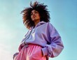 © Viktor - Stylish black woman models trendy modern activewear. Wears fashionable lavender hoodie, pink pants. Photo displays confident fashion in activewear. Outdoor shot against clear sky. Outfit trendy,