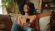 © Seno - Joyful young black woman holding smart phone device sitting on sofa at home