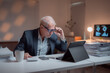 © MINAE - Senior businessman adjusting his glasses and working late at night in office using digital tablet with stacks of paperwork and computer showing graphs on the desk