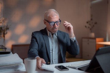 Wall Mural - Focused mature businessman sitting at his desk working late in the office, adjusting his glasses while grabbing a coffee mug, with a pile of paperwork and a tablet in front of him