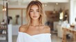 © musa - Attractive young woman with blonde hair and blue eyes, wearing an off-the-shoulder white top, poses indoors.