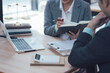 © Kritdanai - Legal Consultation: A legal professional, dressed in a smart suit, carefully reviews a case file with a client. They are seated in a well-lit office with a laptop, calculator, and law books visible.
