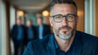 © BROPRADANA - Confident businessman hallway portrait, colleagues blurred background, corporate success