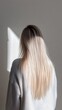 © Sergey - Rear view of a young woman with long, silky blonde hair, standing indoors against a neutral backdrop, softly lit by natural light.