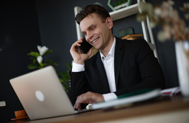 A young man in a black suit smiles while talking on the phone and working on a laptop in a modern office. The workspace includes plants, creating a productive and professional environment.