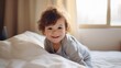 © Iona - A cheerful child with tousled hair beams joyfully from atop white sheets, capturing the essence of simple childhood happiness in a cozy bedroom setting.