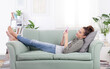 © amedeoemaja - Young relaxed smiling woman lying on a cozy couch in a casual outfit, holding a smartphone and browsing messages, social media, and digital content