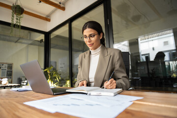 Wall Mural - Middle eastern woman using computer for work online, writing notes, watching webinar. Focused professional latin hispanic financial specialist business lady working concentrated on laptop pc in office