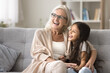 © fizkes - In warm embraces of granny. Supportive senior grandmother hug beloved little preteen girl grandkid sit on sofa look at distance chat dream together plan future trip on vacation create idea of new game
